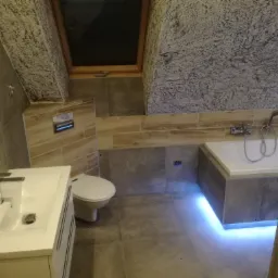 Nowoczesna łazienka na poddaszu z oknem dachowym, szarą glazurą, białą armaturą i podświetleniem LED wzdłuż wanny.