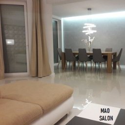 MAO Magdalena Owczarek - Jasne, przestronne wnętrze salonu z jadalnią. Parkiet odbija światło. W tle stół z szarymi krzesłami, nad nim designerska lampa. Ściana w odcieniach szarości z subtelnym oświetleniem LED. Z lewej...