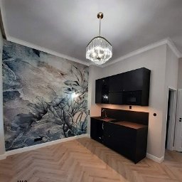 MAO Magdalena Owczarek - Wnętrze małej kuchni w kamienicy z czarnymi szafkami, drewnianym blatem, parkietem ułożonym w jodełkę i tapetą z motywem kwiatowym w odcieniach szarości i błękitu, oświetlone kryształowym żyrandolem.