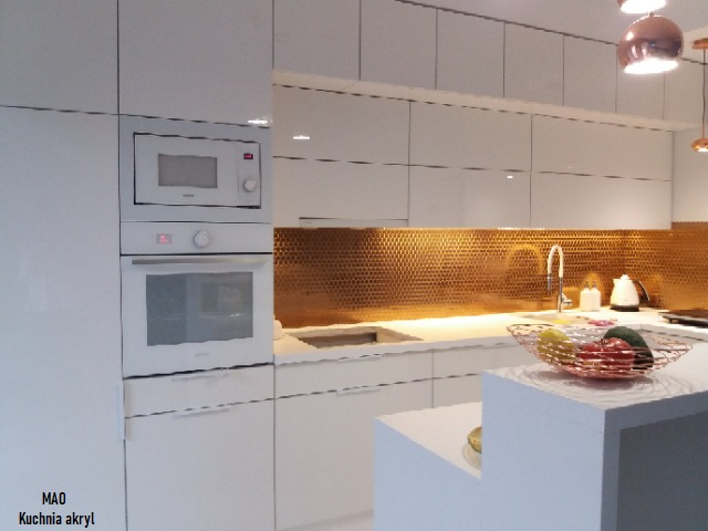 Biała kuchnia akrylowa z wbudowanym piekarnikiem i mikrofalówką, miedziana mozaika na ścianie nad blatem, owocowa misa na wyspie.