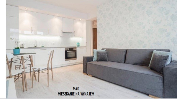 Jasne wnętrze mieszkania z aneksem kuchennym, szarą sofą i ścianą w delikatny wzór kwiatowy. Białe, lakierowane szafki kuchenne, stół z krzesłami w stylu skandynawskim i drewniana podłoga.