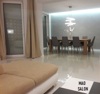 Jasne, przestronne wnętrze salonu z jadalnią. Parkiet odbija światło. W tle stół z szarymi krzesłami, nad nim designerska lampa. Ściana w odcieniach szarości z subtelnym oświetleniem LED. Z lewej...