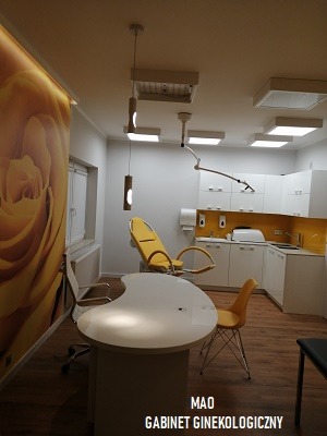 Jasne wnętrze gabinetu lekarskiego z żółtym fotelem zabiegowym, białym biurkiem o nieregularnym kształcie, szafkami w tle i tapetą z motywem żółtej róży na ścianie.