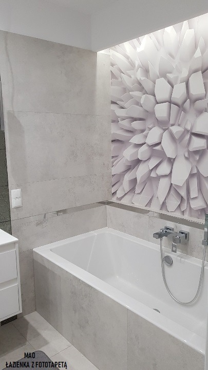 Nowoczesna łazienka z białą wanną obudowaną szarymi płytkami imitującymi beton, akcentująca ściana z fototapetą 3D w geometryczny wzór.