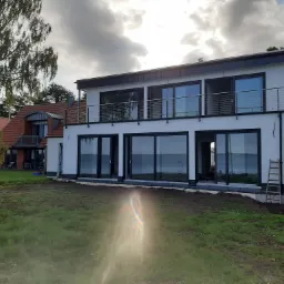 Nowoczesny, dwupiętrowy dom z dużymi przeszkleniami, czarnymi ramami okiennymi i balkonem z metalową balustradą. Na trawniku przed domem stoi drabina.