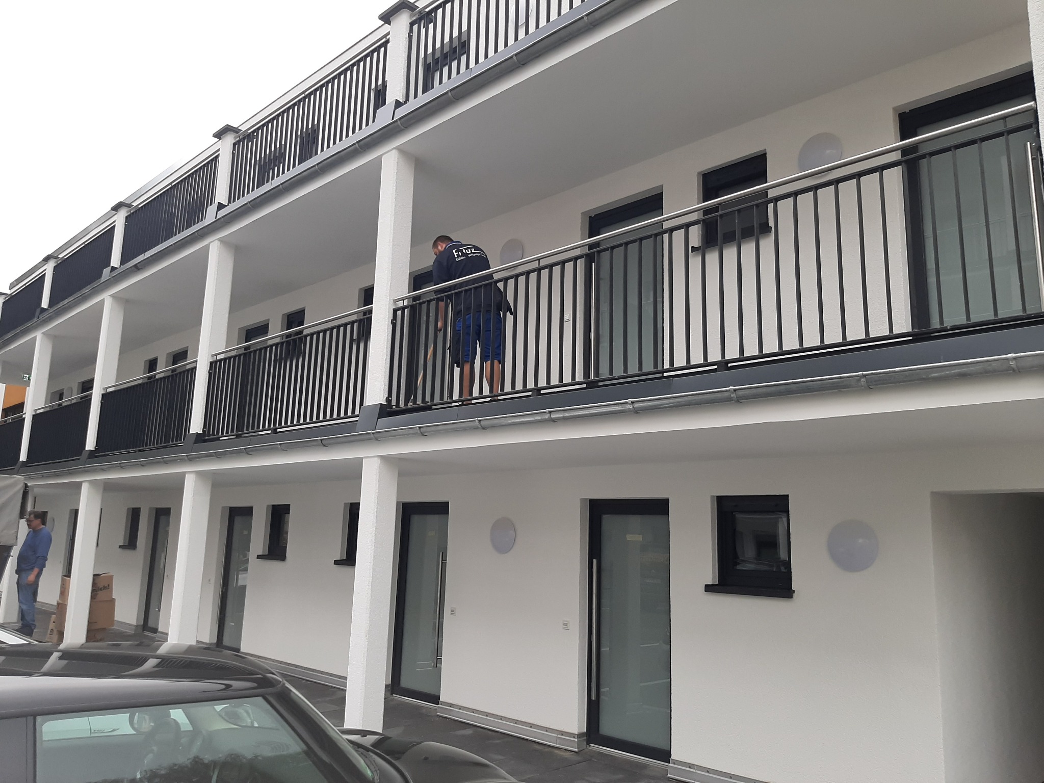 Montażysta na balkonie budynku wielorodzinnego, instaluje zabezpieczenie okienne, widoczne czarne ramy okienne i balustrady balkonowe.