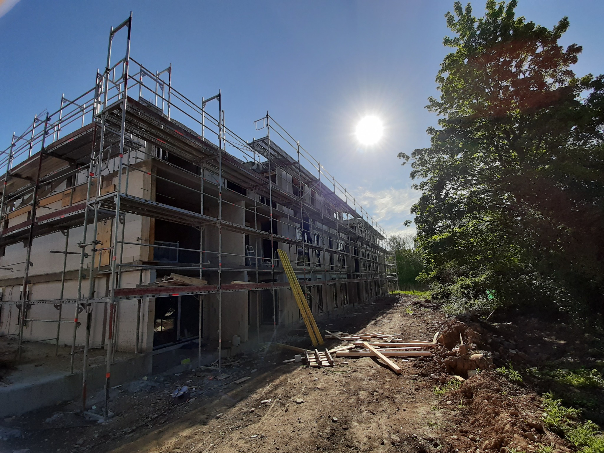 Budynek wielorodzinny w trakcie budowy, z widocznymi rusztowaniami, niezamontowanymi oknami i elementami drewnianymi na placu budowy, w słoneczny dzień.