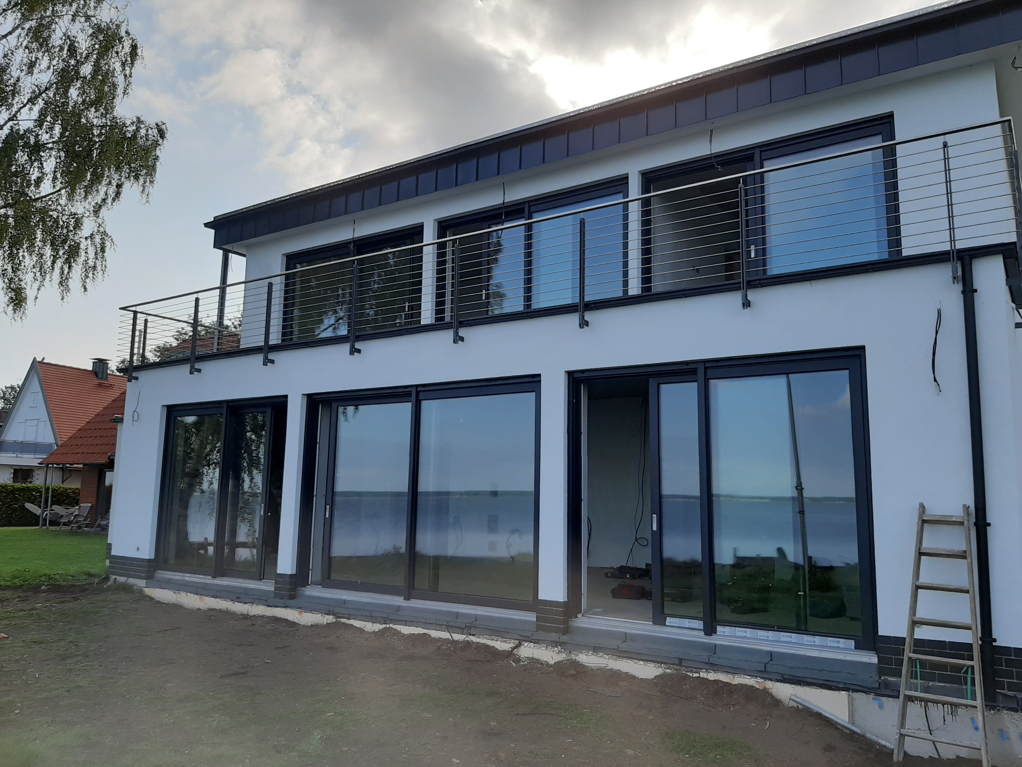 Nowoczesny dom z białymi ścianami i dużymi przesuwnymi oknami z czarnymi ramami, widok na jezioro, balkon z metalową balustradą, drabina oparta o ścianę.