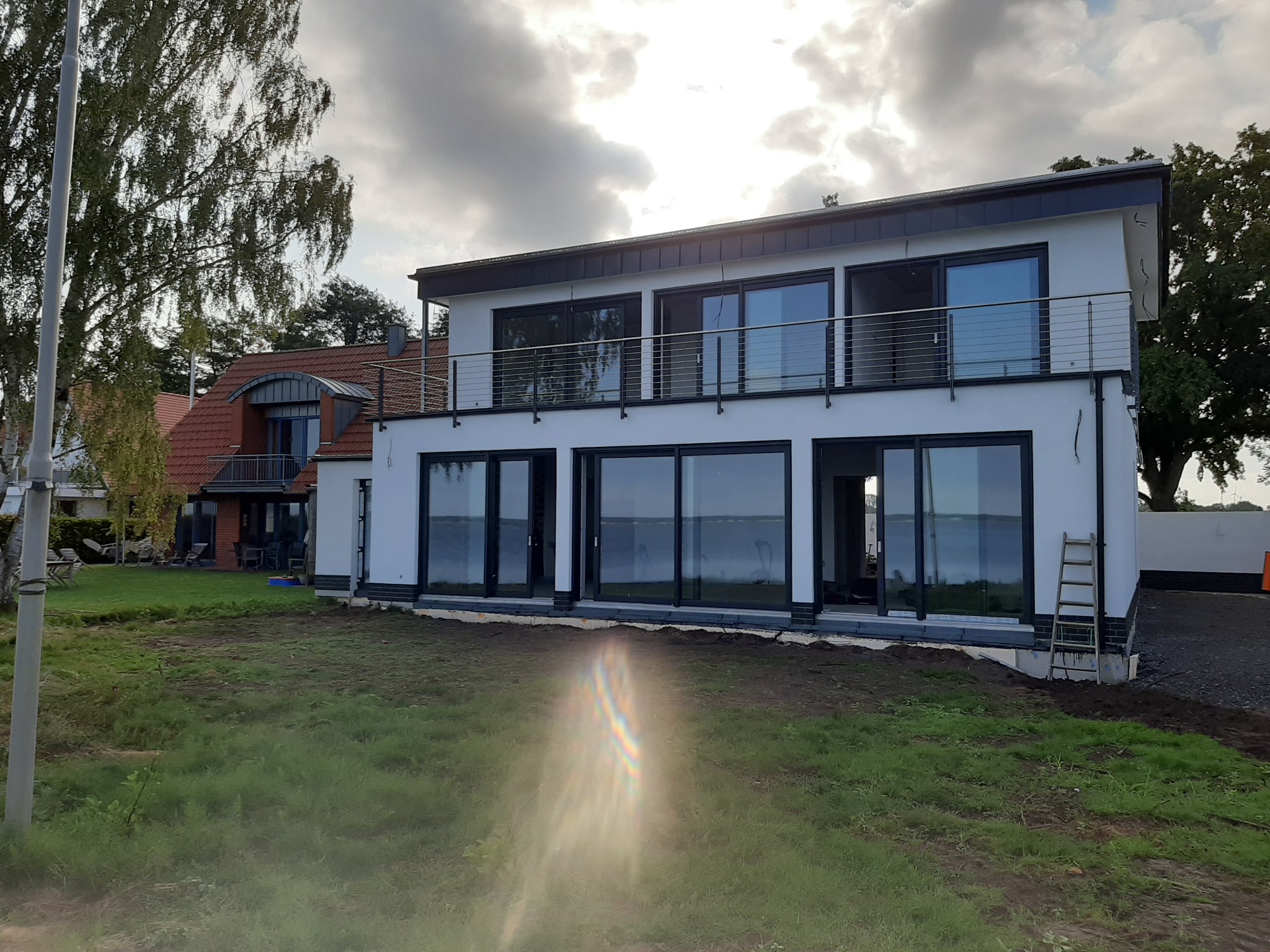 Nowoczesny, dwupiętrowy dom z dużymi przeszkleniami, czarnymi ramami okiennymi i balkonem z metalową balustradą. Na trawniku przed domem stoi drabina.