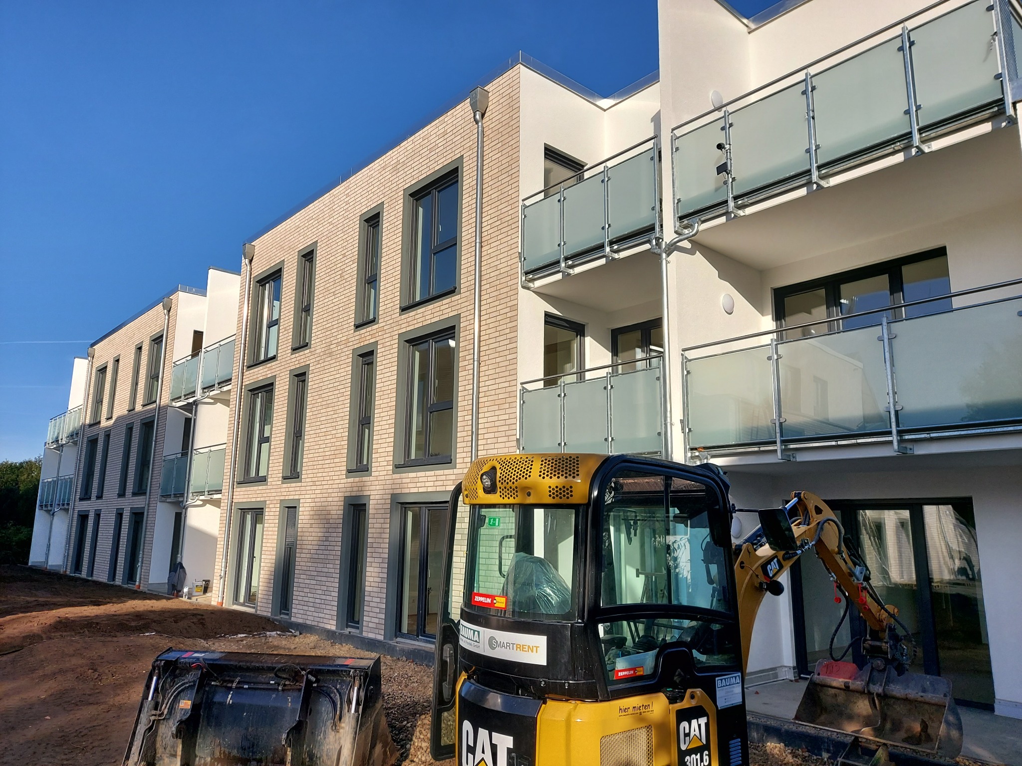 Nowo wybudowany budynek wielorodzinny z jasnej cegły, z balkonami z matowego szkła, widoczny z perspektywy żółtej koparki CAT 301.6 na placu budowy w słoneczny dzień.