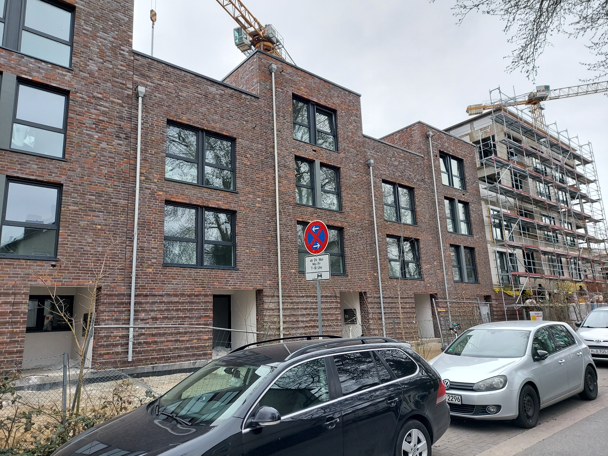 Nowe budynki mieszkalne z czerwonej cegły z czarnymi ramami okiennymi, częściowo w trakcie budowy, z widocznymi żurawiami budowlanymi i rusztowaniami, zaparkowane samochody na pierwszym planie.