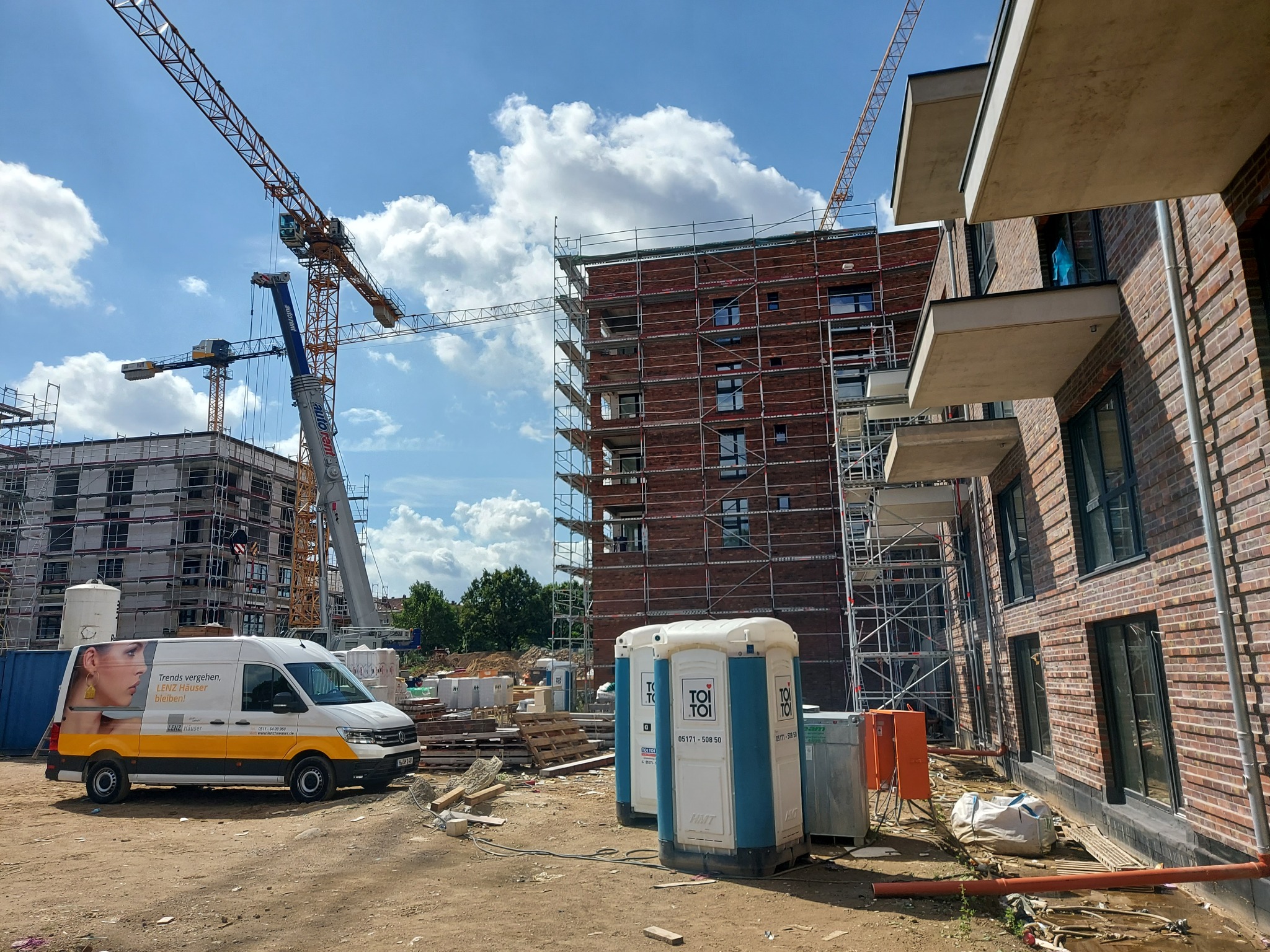 Budowa budynku mieszkalnego z cegły, widoczne rusztowania, żurawie budowlane, przenośne toalety TOI TOI i samochód dostawczy z reklamą Lenz Häuser na placu budowy.