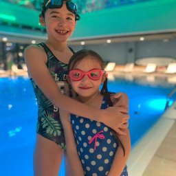 Michał Czerkas - Dwie uśmiechnięte dziewczynki w strojach kąpielowych i okularach pływackich stoją przy basenie z turkusową wodą, starsza obejmuje młodszą ramieniem, w tle leżaki.