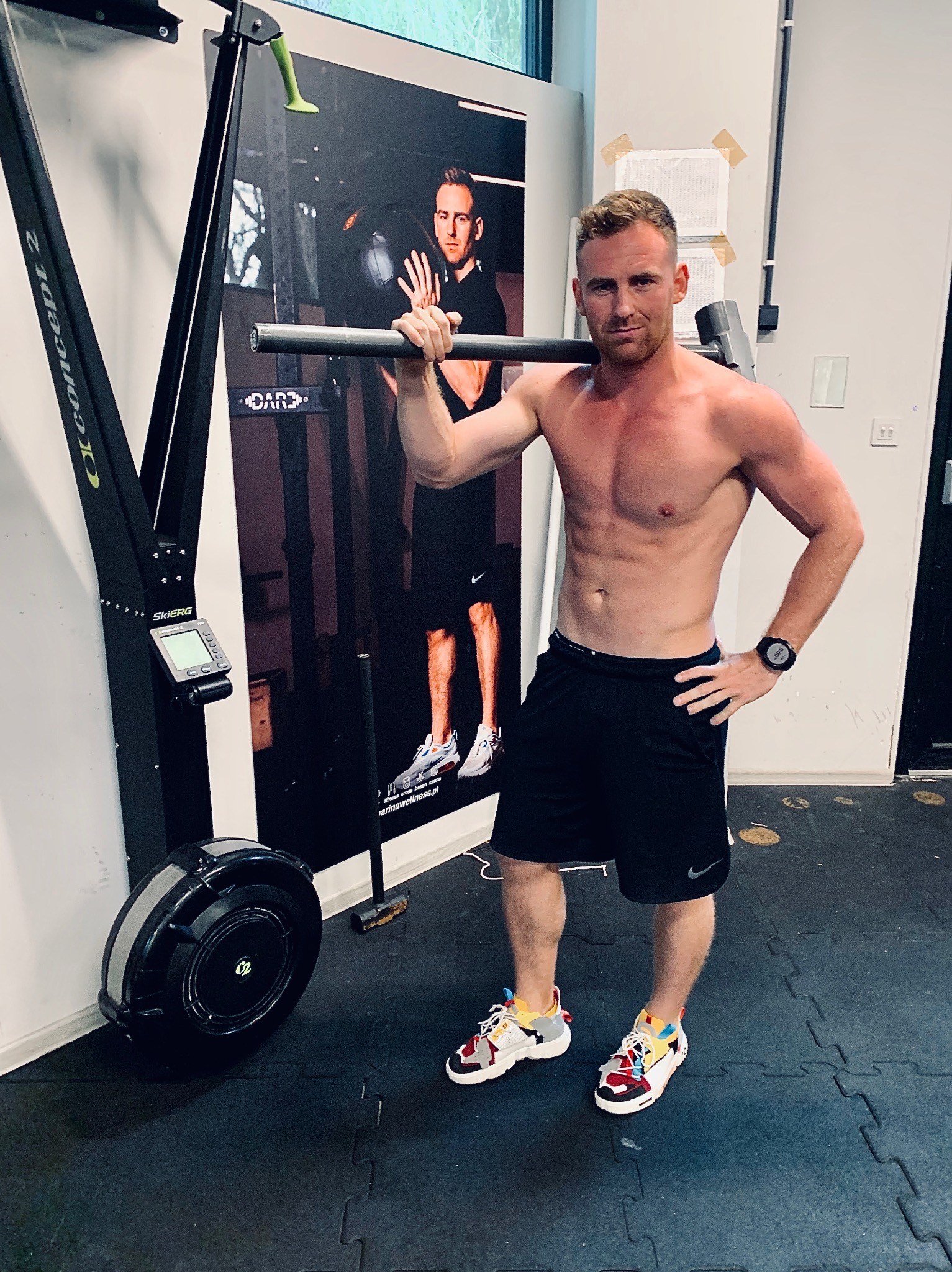 Mężczyzna bez koszulki z sztangą na ramionach, stojący w siłowni obok ergometru narciarskiego i plakatu z innym mężczyzną podczas treningu z piłką lekarską.