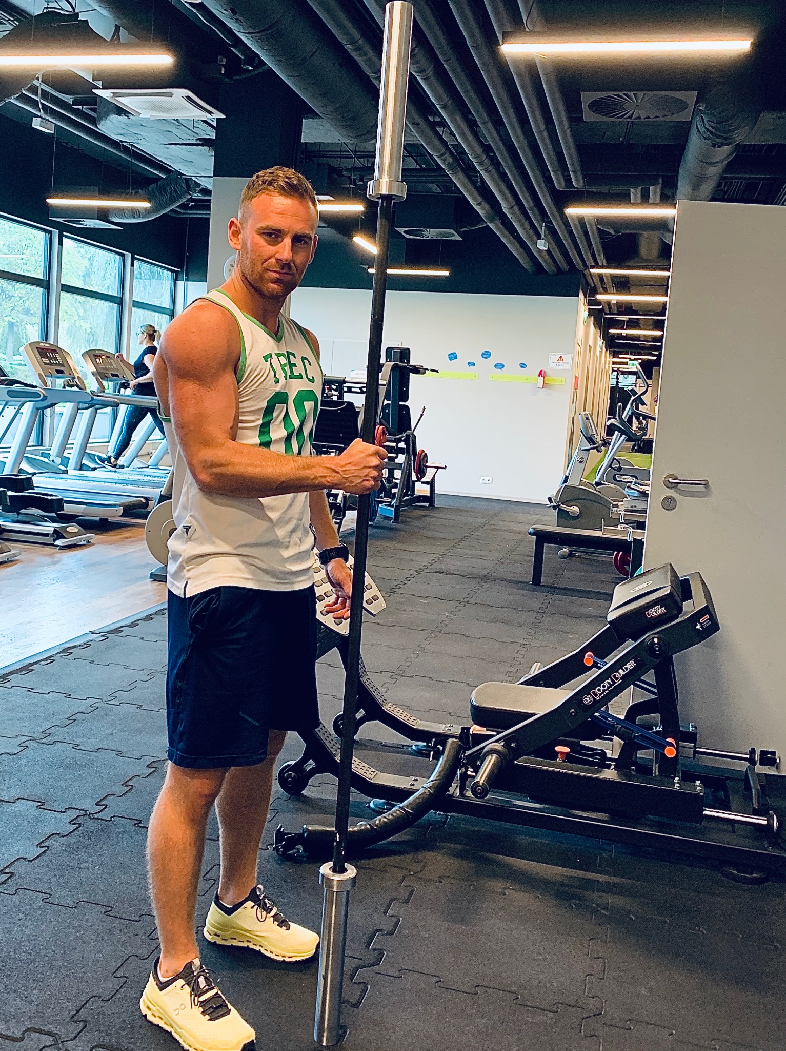 Mężczyzna w sportowym stroju trzyma sztangę na tle siłowni z bieżniami i sprzętem do ćwiczeń, widoczny mocno zarysowany biceps.