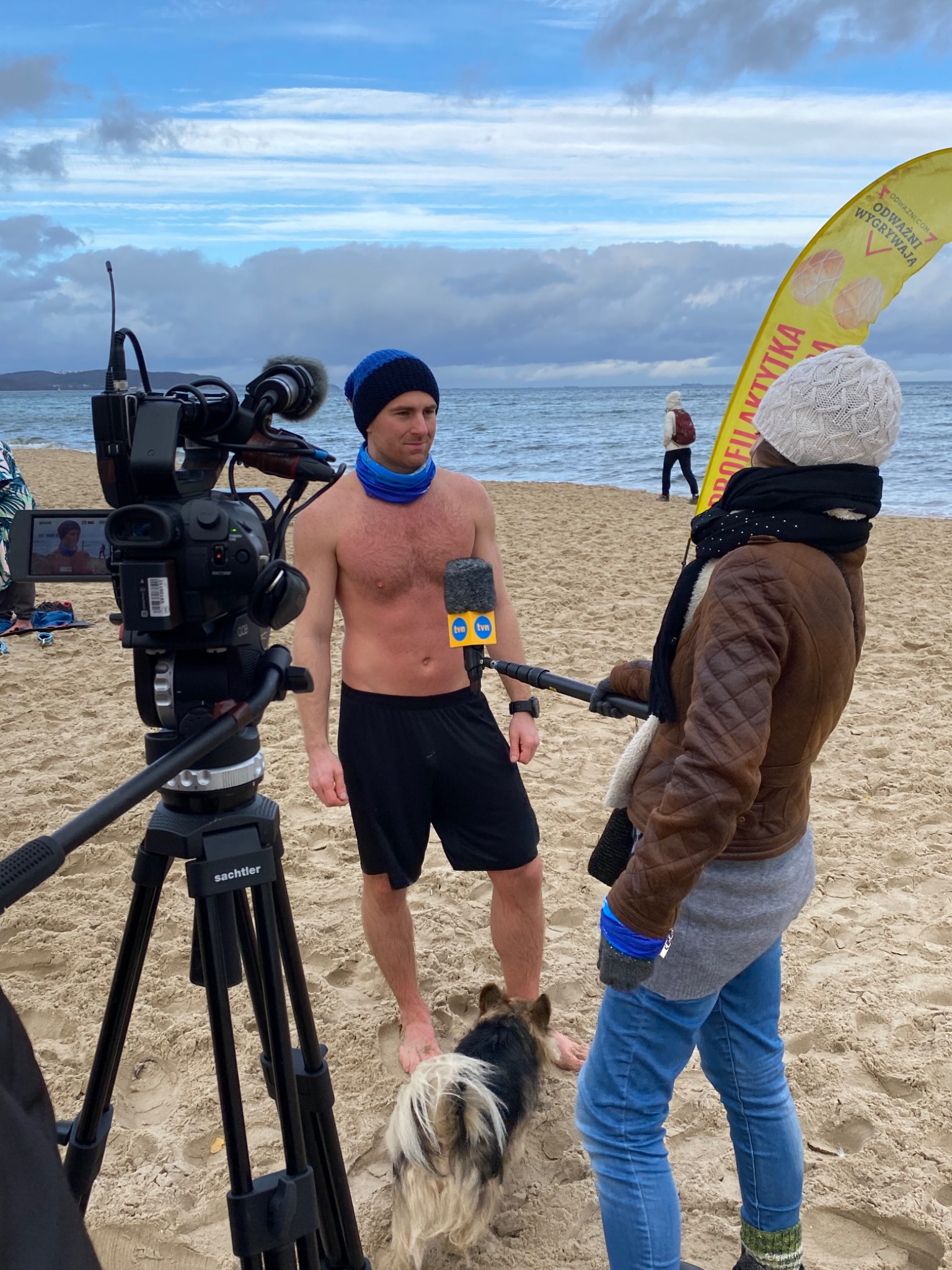 Mężczyzna bez koszulki w czapce i szaliku, trzymający mikrofon TVN, stoi na plaży podczas relacji na żywo; obok pies i operator kamery.