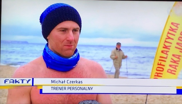 Relacja telewizyjna z plaży: mężczyzna w czapce i chuście, z widoczną częścią torsu, w tle morze i osoba uprawiająca nordic walking, obok żółty baner z napisem.