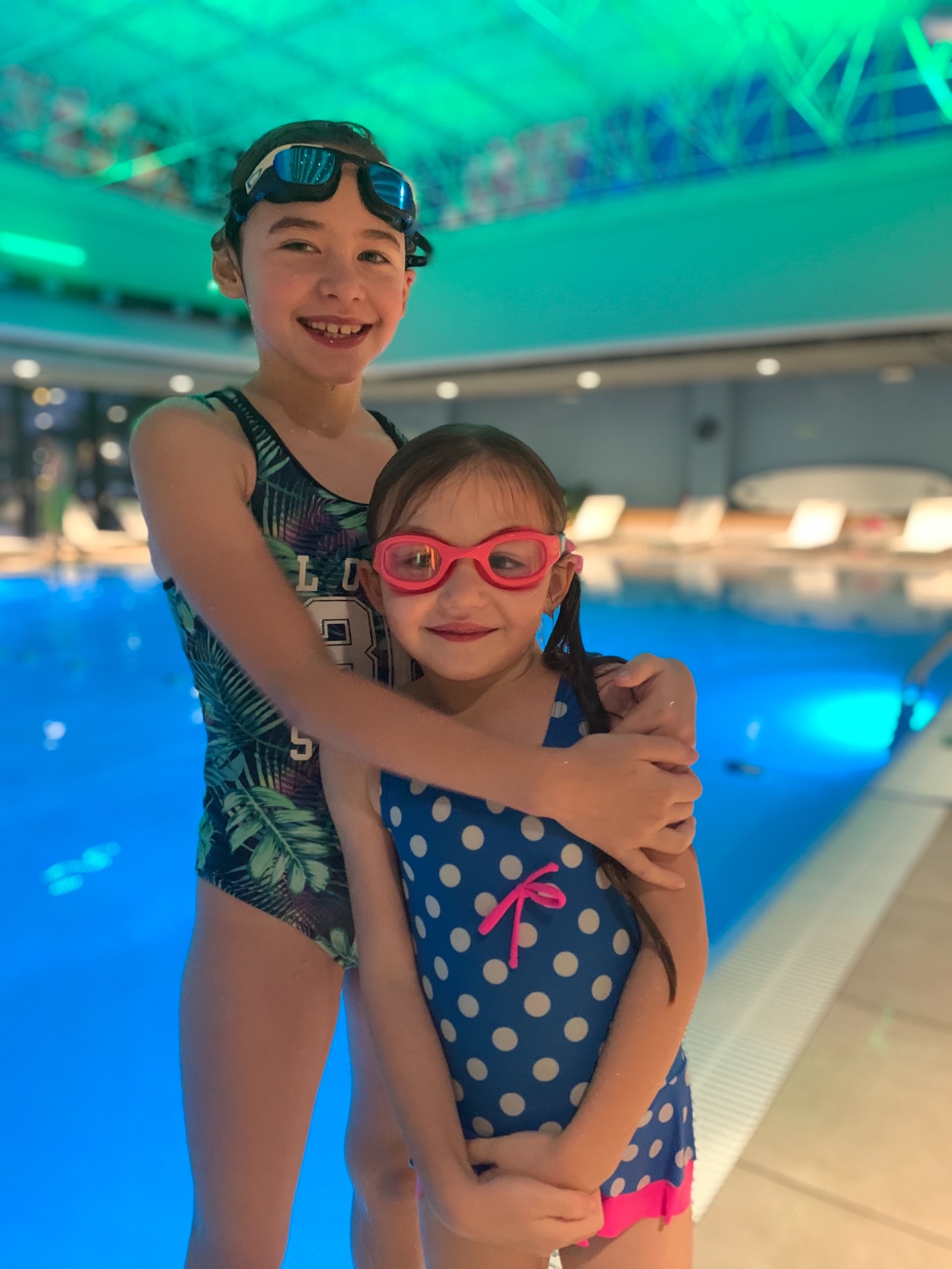 Dwie uśmiechnięte dziewczynki w strojach kąpielowych i okularach pływackich stoją przy basenie z turkusową wodą, starsza obejmuje młodszą ramieniem, w tle leżaki.