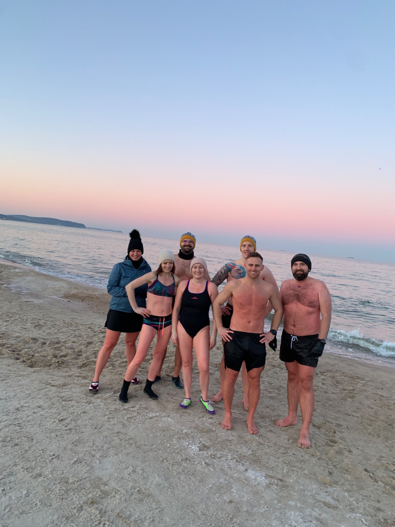 Grupa sześciu osób w strojach kąpielowych i czapkach stoi na piaszczystej plaży nad morzem o zachodzie słońca, z widocznymi falami i odległym brzegiem.