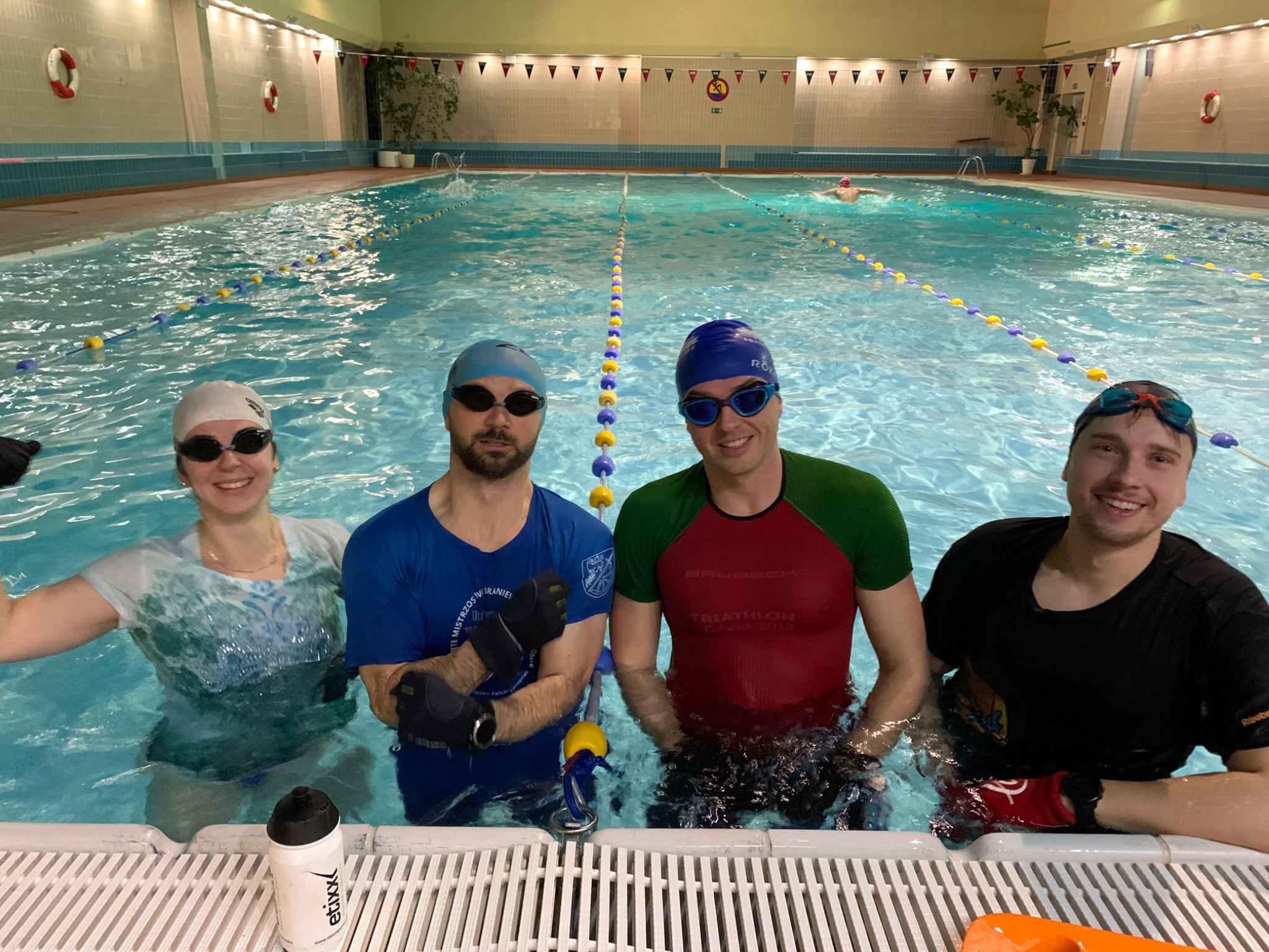 Czterech uśmiechniętych pływaków w czepkach i okularach stoi w basenie, w tle tor z bojkami i osoba pływająca kraulem, na pierwszym planie bidon z napojem izotonicznym.