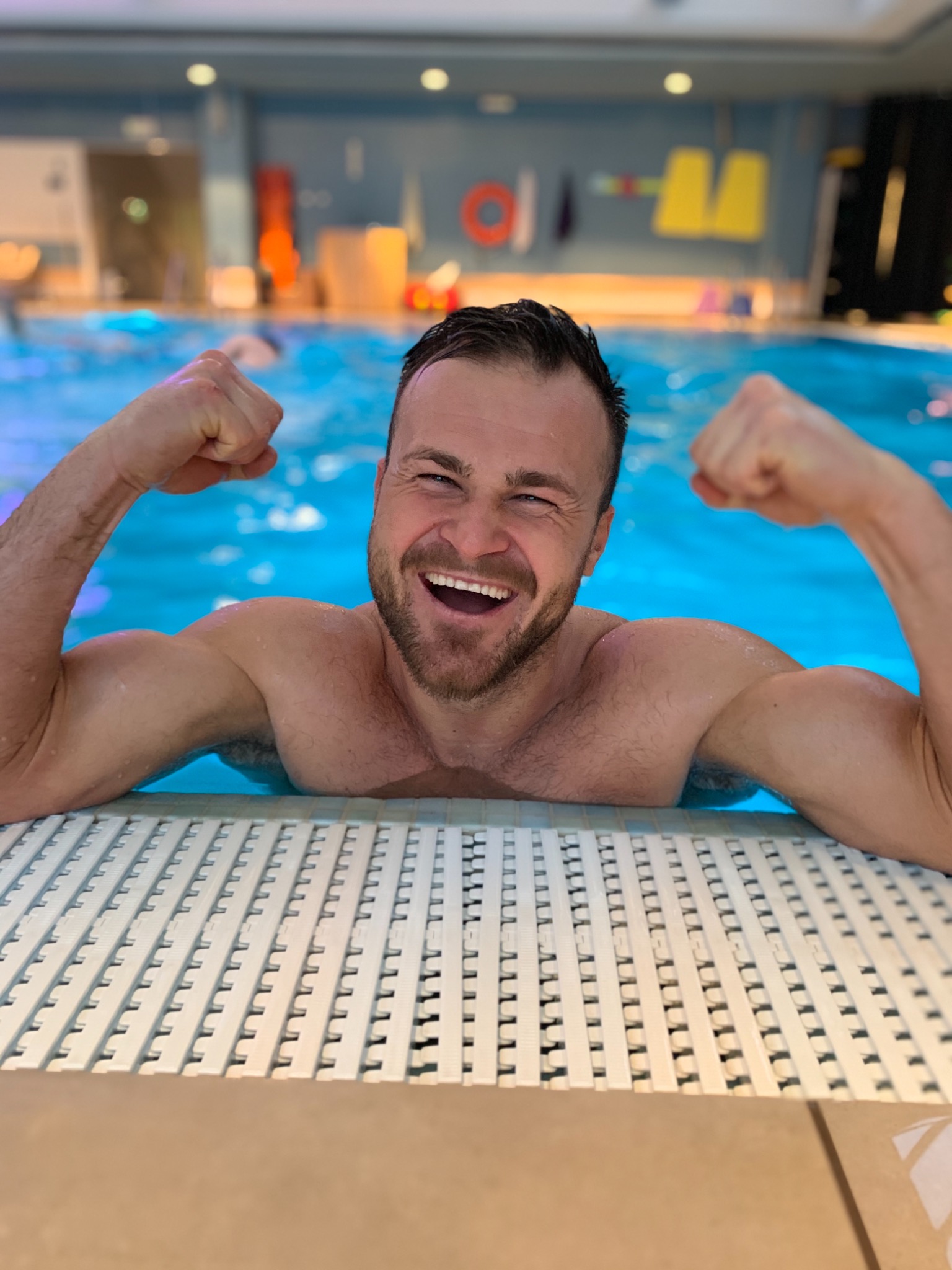Uśmiechnięty mężczyzna z zaciśniętymi pięściami w basenie, w tle widoczne akcesoria basenowe i niebieska woda.