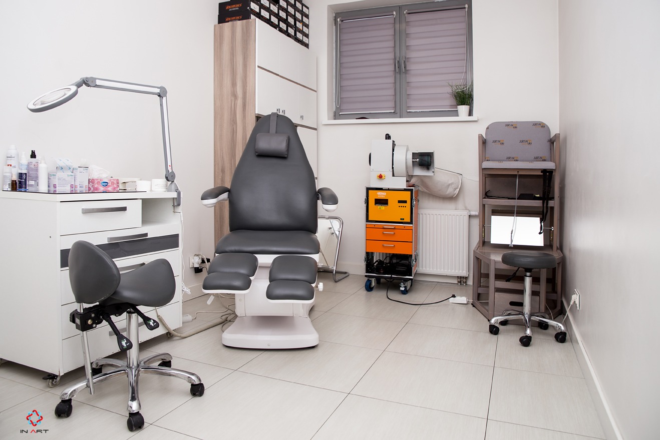 Nowoczesny gabinet kosmetyczny z fotelem do zabiegów, lampą powiększającą, szafką z kosmetykami, urządzeniem do sterylizacji i krzesłem na kółkach.