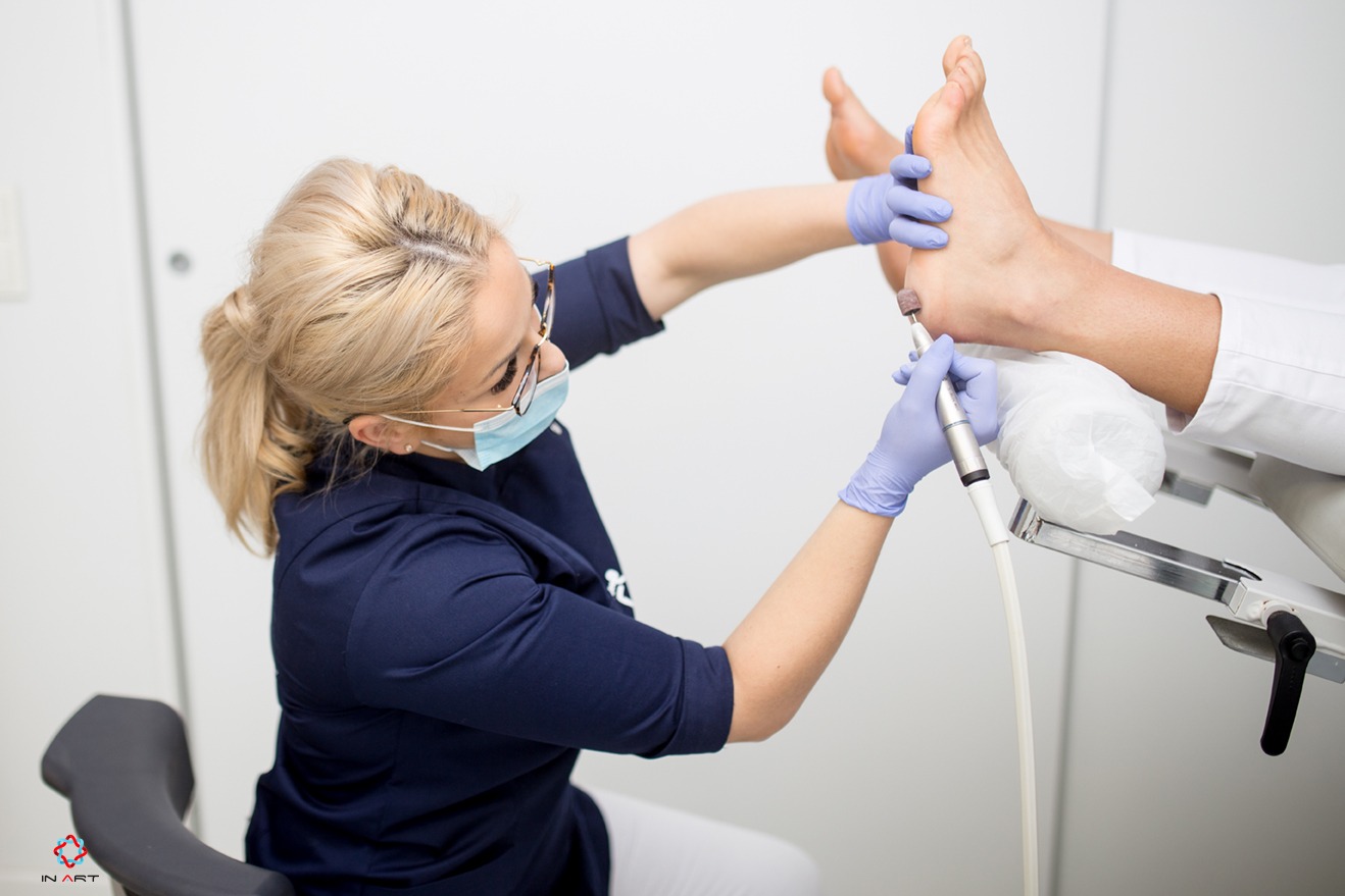 Podolog w niebieskiej bluzie i rękawiczkach, w masce na twarzy, szlifuje piętę pacjenta leżącego na fotelu za pomocą frezarki.