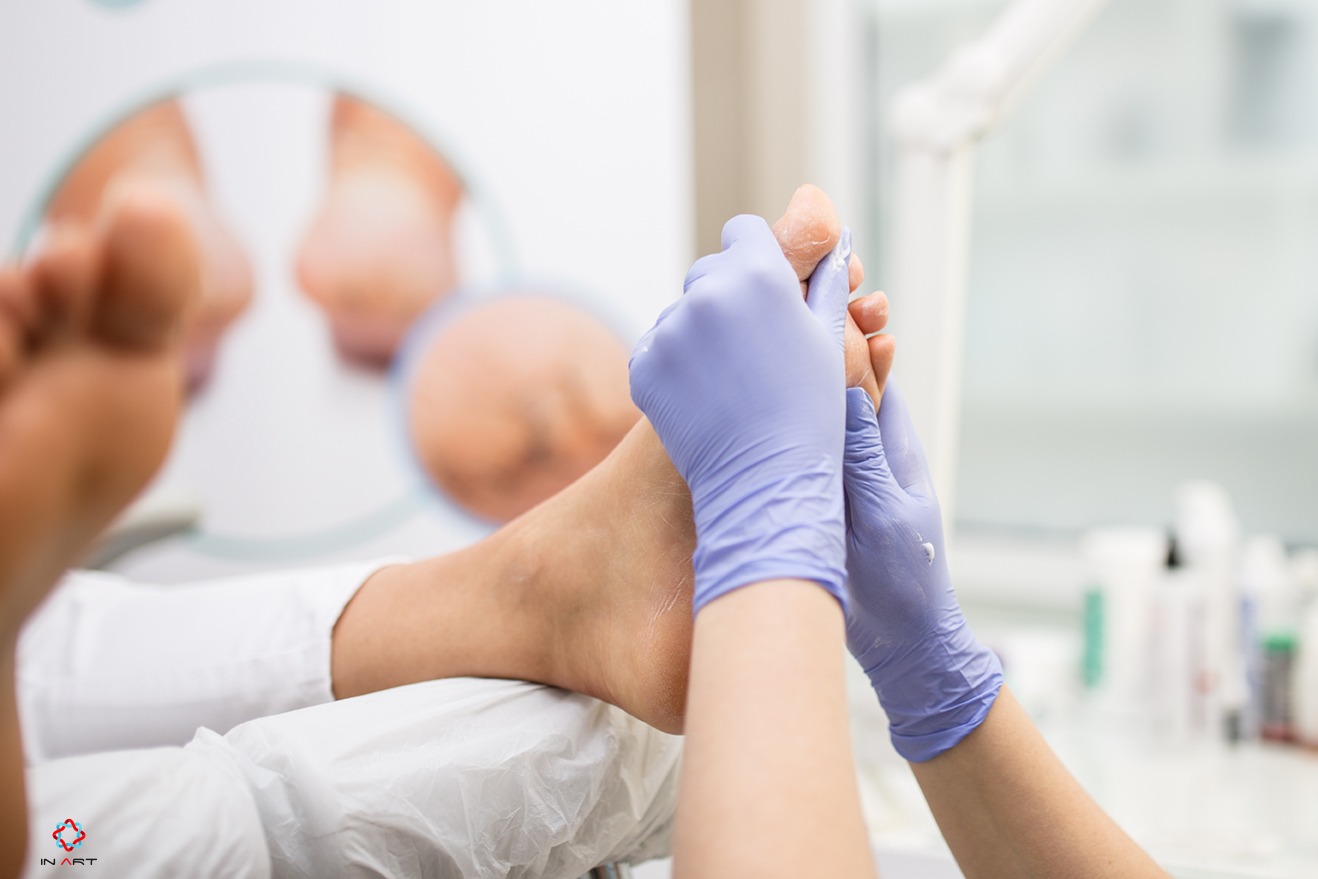 Pedicure w trakcie wykonywania: dłoń w lateksowej rękawiczce nakłada krem na stopę klienta leżącego na białym podkładzie, w tle widoczny sprzęt kosmetyczny i stopy klienta w okrągłym lustrze.