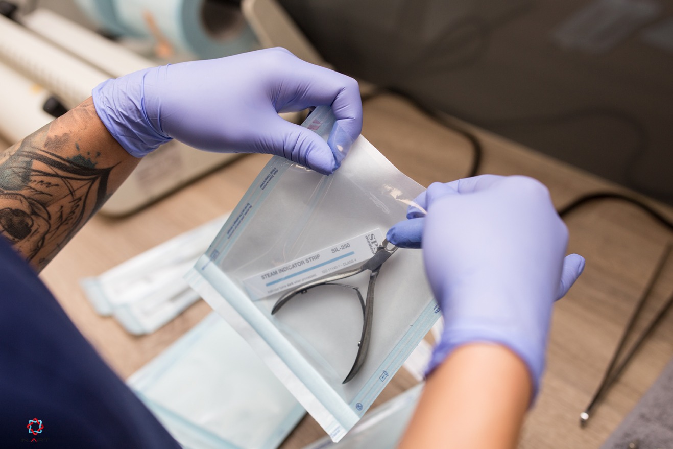 Otwieranie sterylnego opakowania z cążkami do skórek, dłonie w niebieskich rękawiczkach, widoczny tatuaż na ramieniu.