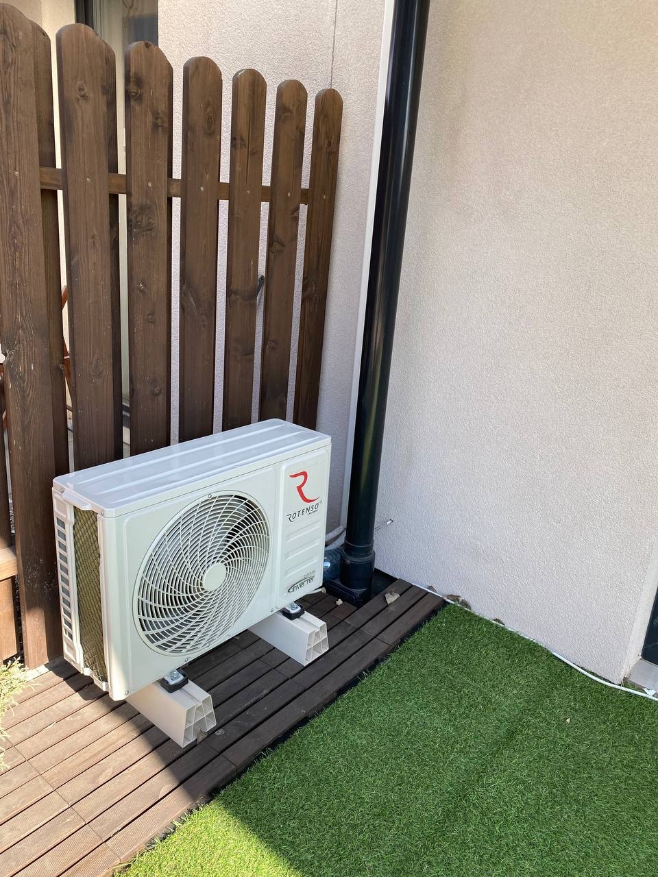 Biała jednostka zewnętrzna klimatyzatora Rotenso Elis 3.5kW zamontowana na plastikowych wspornikach na drewnianym podeście obok sztucznej trawy i brązowego płotu.