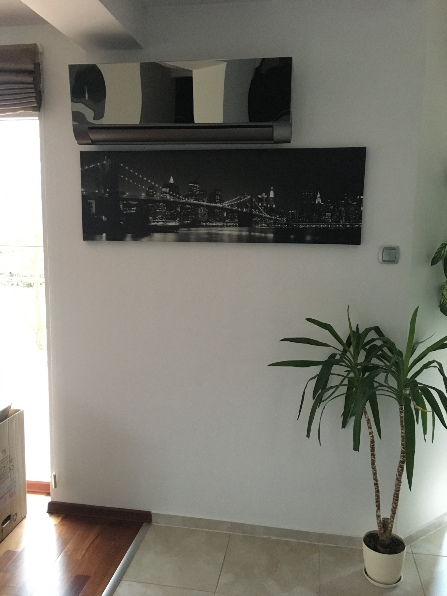 Zainstalowany klimatyzator Rotenso Versu Mirror 5kW w nowoczesnym wnętrzu z obrazem panoramy miasta i rośliną doniczkową.