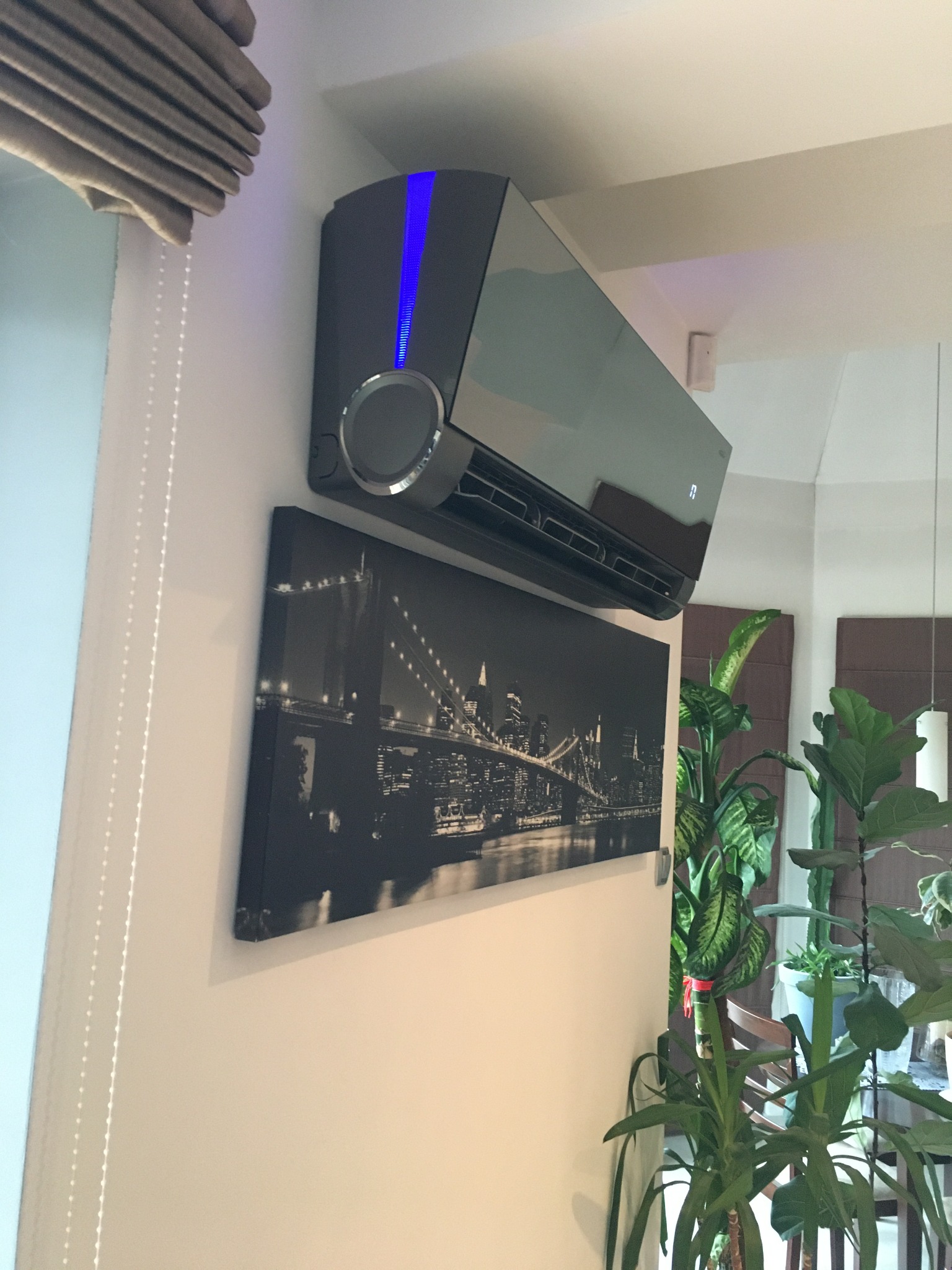 Nowoczesny klimatyzator Rotenso Versu Mirror 5kW zamontowany na ścianie nad czarno-białym obrazem panoramy miasta, widoczne rośliny doniczkowe i fragment żaluzji.