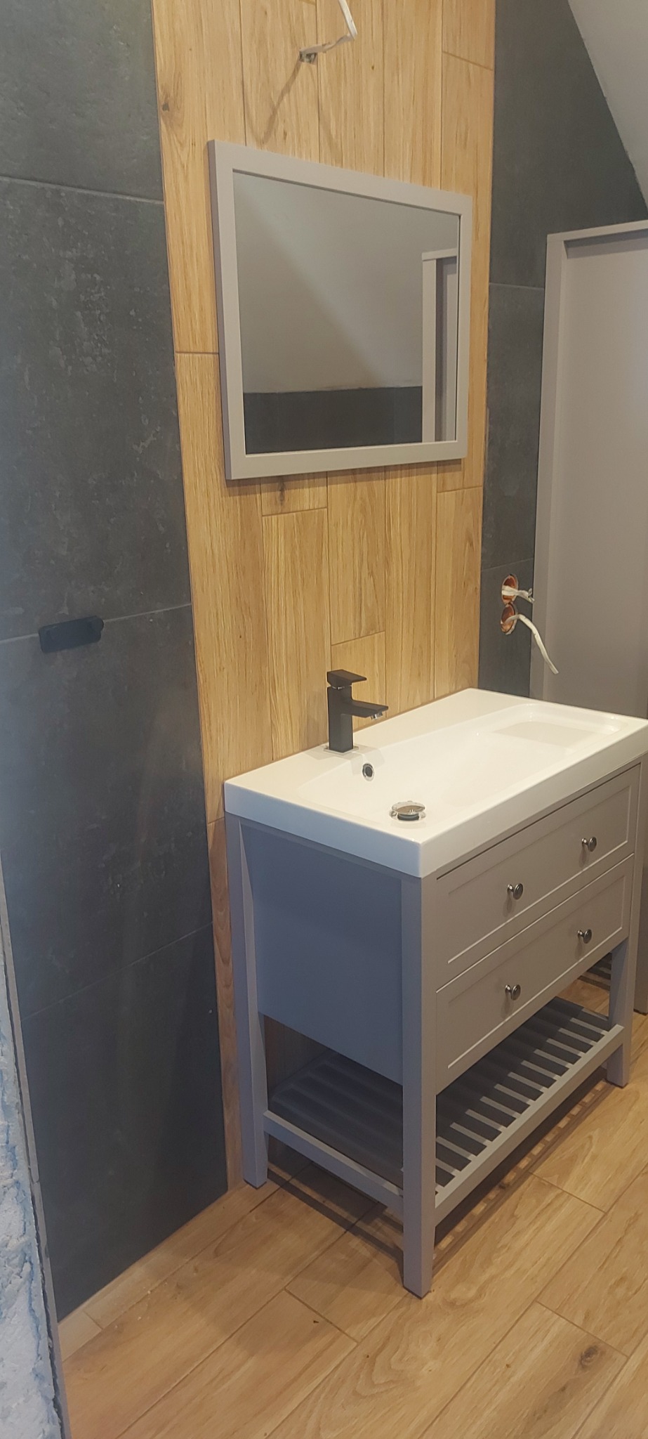 Szara szafka pod umywalkę z czarną baterią, lustro w szarej ramie na ścianie wyłożonej drewnopodobnymi panelami, fragment ściany z ciemnymi płytkami, widoczne niedokończone podłączenie elektryczne.