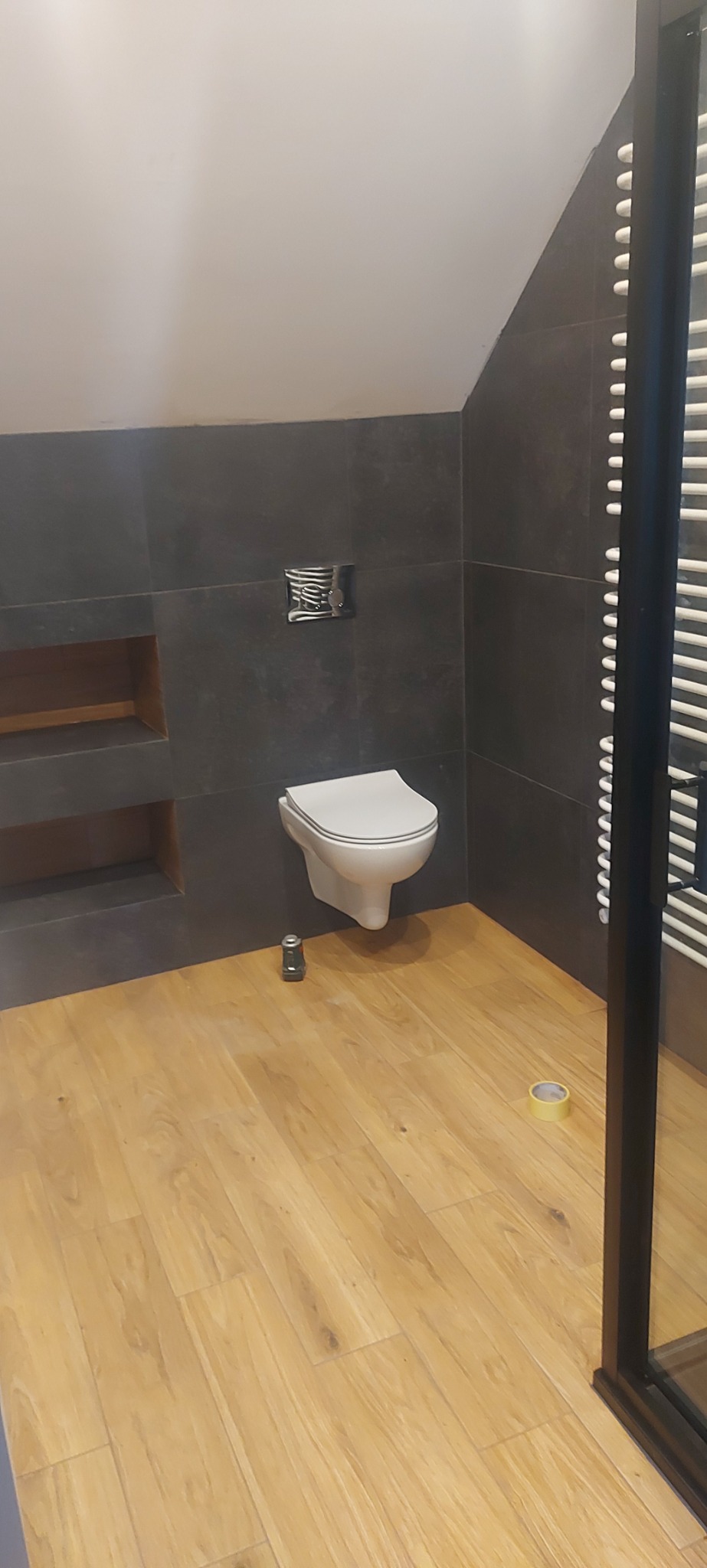 Nowoczesna łazienka na poddaszu z szarymi płytkami, drewnopodobną podłogą i wiszącą toaletą. Widoczny grzejnik drabinkowy, puszka i taśma malarska.