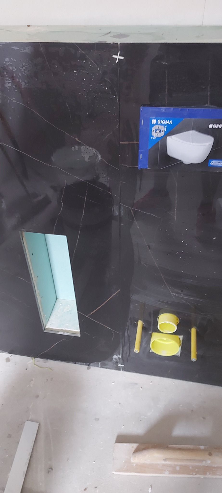 Częściowo wyłożona czarnymi płytkami ściana łazienki z otworem na instalację, widoczne elementy montażowe i opakowanie produktu Sigma/Geberit.