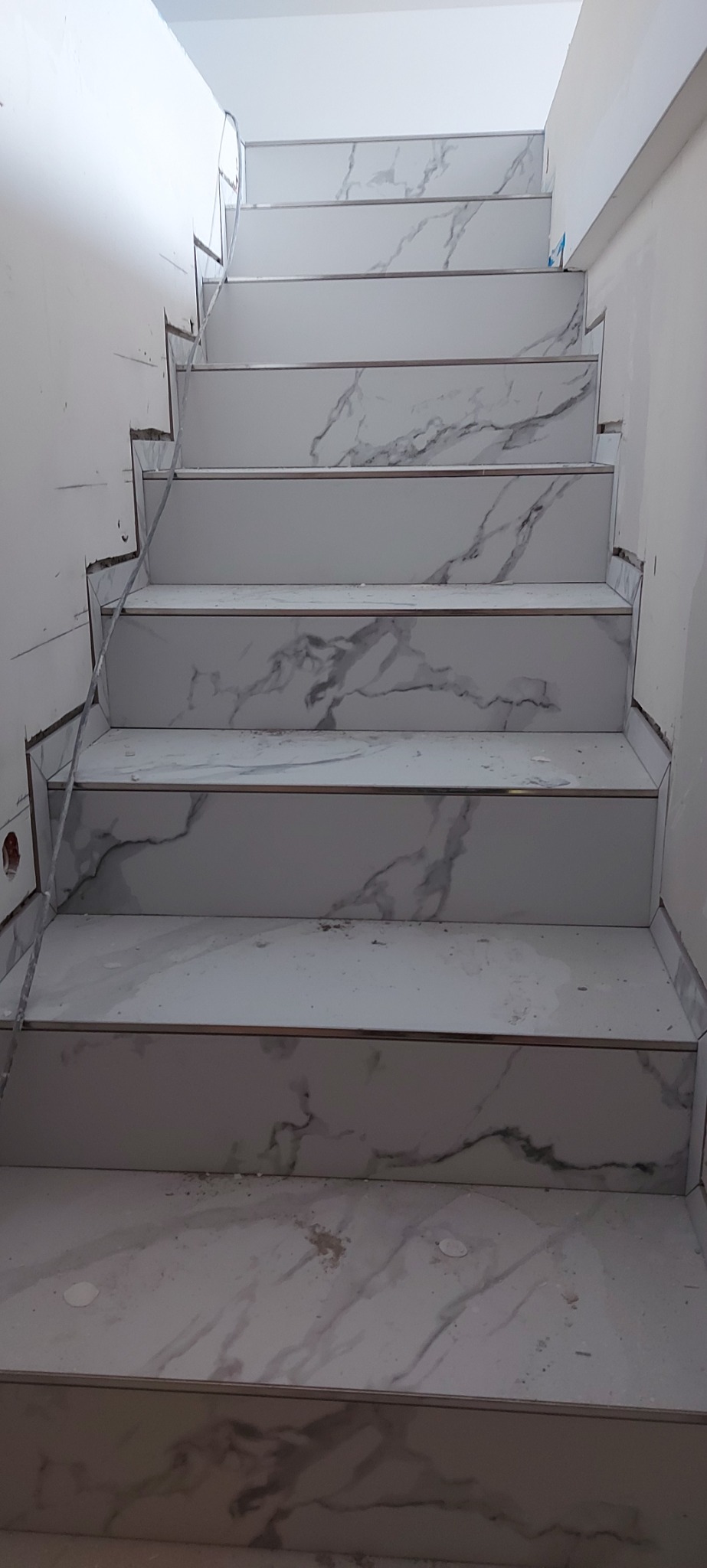 Schody wyłożone białymi płytkami imitującymi marmur, z metalowymi listwami krawędziowymi, w trakcie prac wykończeniowych.