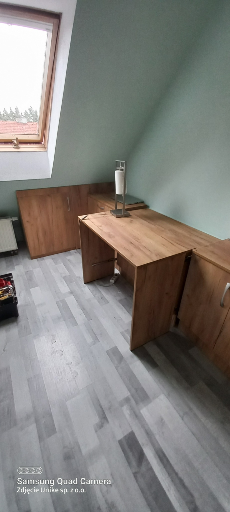 Drewniane meble do zabudowy pod skosem na poddaszu, z biurkiem i szafkami, w trakcie montażu, widoczny grzejnik i skrzynka z narzędziami.