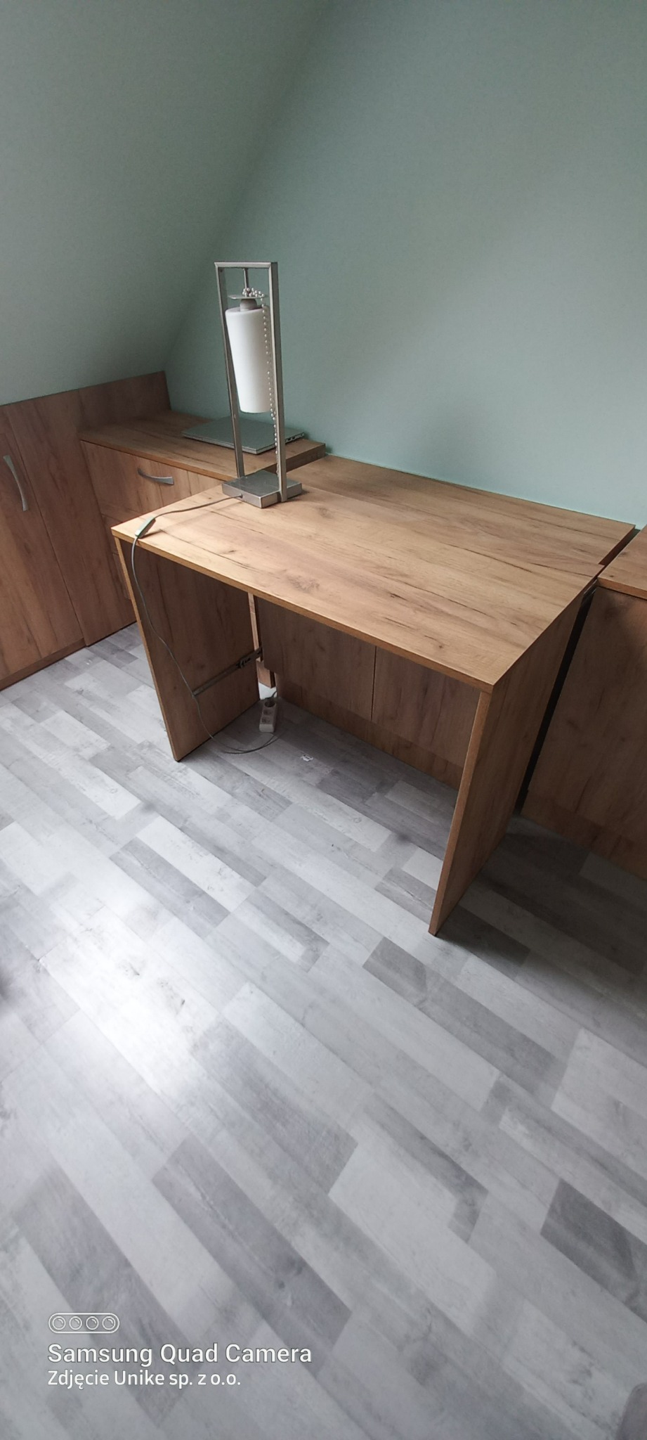 Drewniany blat biurka z szafkami w zabudowie, oświetlony lampą biurkową na tle jasnej ściany i podłogi w panelach.