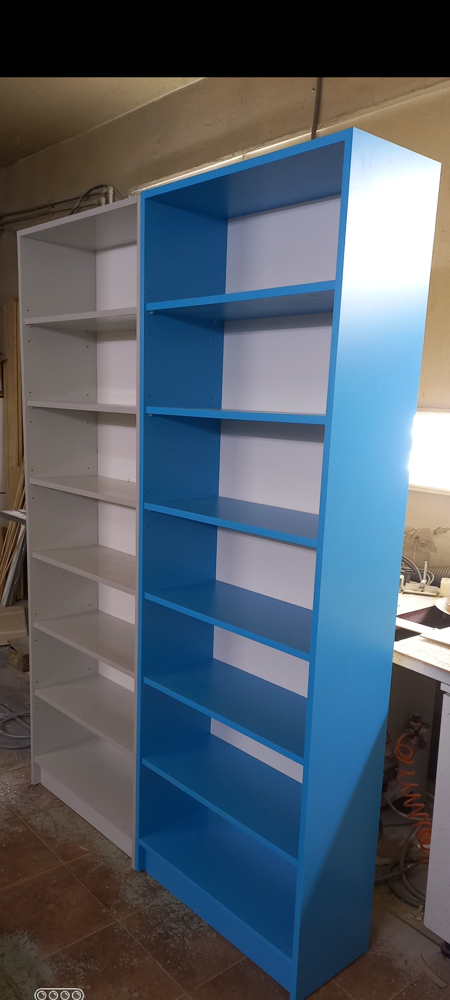 Dwa regały na książki, jeden biały, drugi błękitny, stojące obok siebie w warsztacie stolarskim, widoczne ślady obróbki drewna na podłodze.