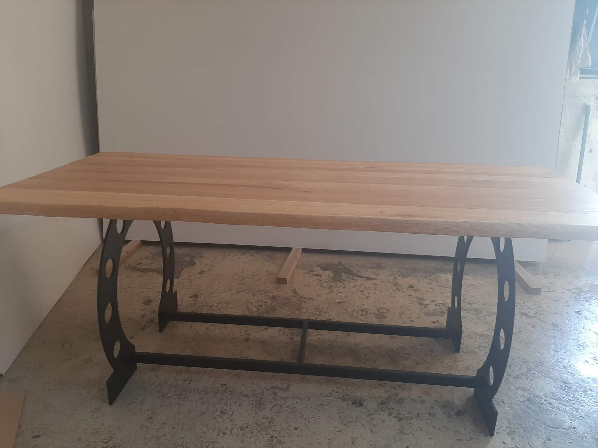 Surowy blat stołu z jasnego drewna, wsparty na czarnej, metalowej konstrukcji z dekoracyjnymi otworami, sfotografowany w warsztatowym otoczeniu.