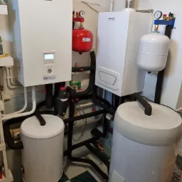 Kompletna instalacja grzewcza w kotłowni: biała jednostka Panasonic z wyświetlaczem, czerwony zbiornik ciśnieniowy, biały piec Brötje z kominem oraz dwa szare zasobniki ciepłej wody, połączone...