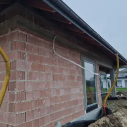 Budynek w trakcie budowy z widocznymi przewodami wentylacyjnymi i rurami wystającymi na zewnątrz, ściany z czerwonej cegły, szara pogoda.