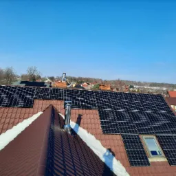 Brązowy dach z panelami fotowoltaicznymi w zimowej scenerii, widoczny komin i okno dachowe, błękitne niebo w tle.