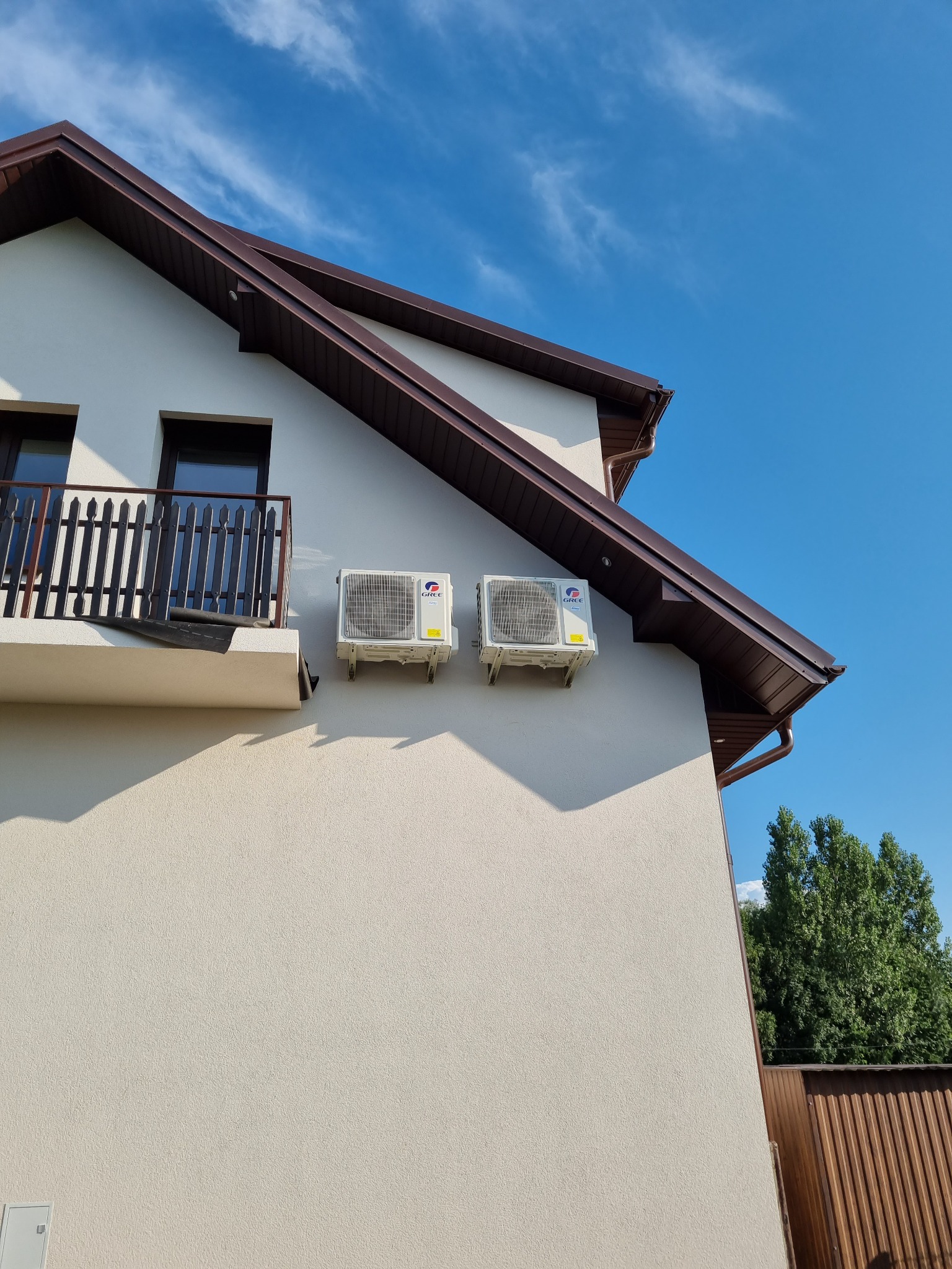 Dwa białe klimatyzatory zamontowane na elewacji jasnego budynku z balkonem, widoczne brązowe elementy dachu i błękitne niebo