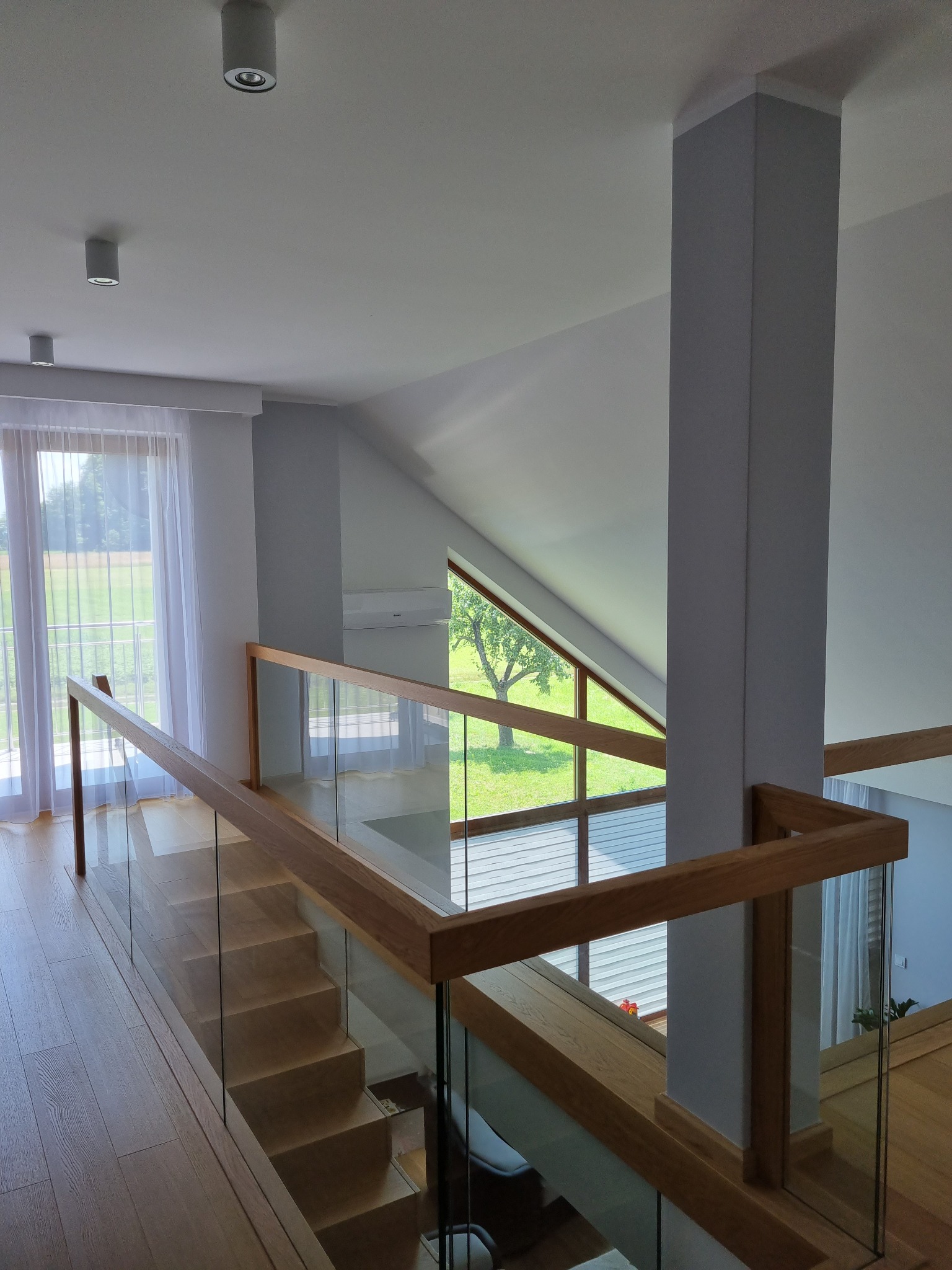 Nowoczesne wnętrze domu z drewnianymi schodami, szklaną balustradą i widokiem na zewnątrz przez okno dachowe, oświetlone punktowymi lampami sufitowymi.