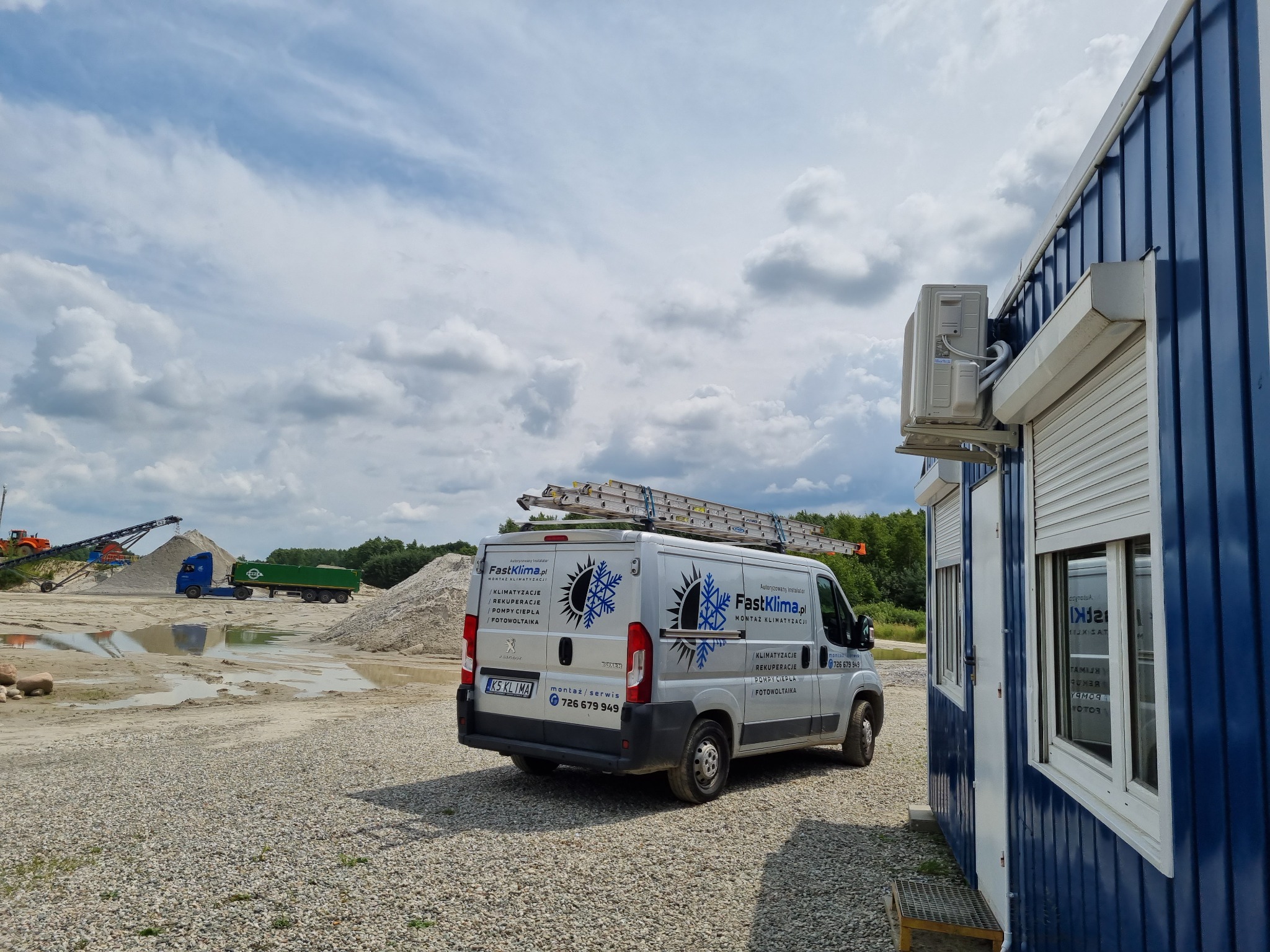 Srebrny samochód dostawczy z logo firmy klimatyzacyjnej i fotowoltaicznej, zaparkowany na żwirowym placu budowy obok niebieskiego kontenera biurowego z zamontowaną jednostką zewnętrzną klimatyzacji...