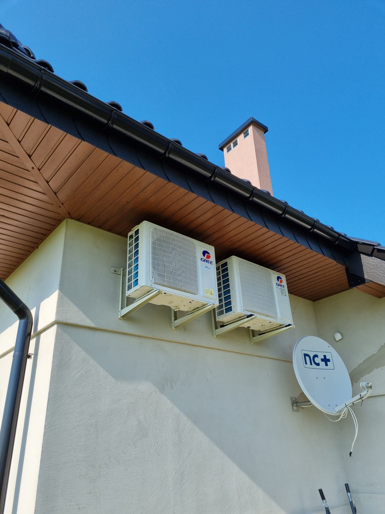 Dwa białe klimatyzatory marki Gree zamontowane na ścianie budynku z jasną elewacją, widoczna antena satelitarna, dach z brązową podbitką i czarną dachówką, błękitne niebo w tle.