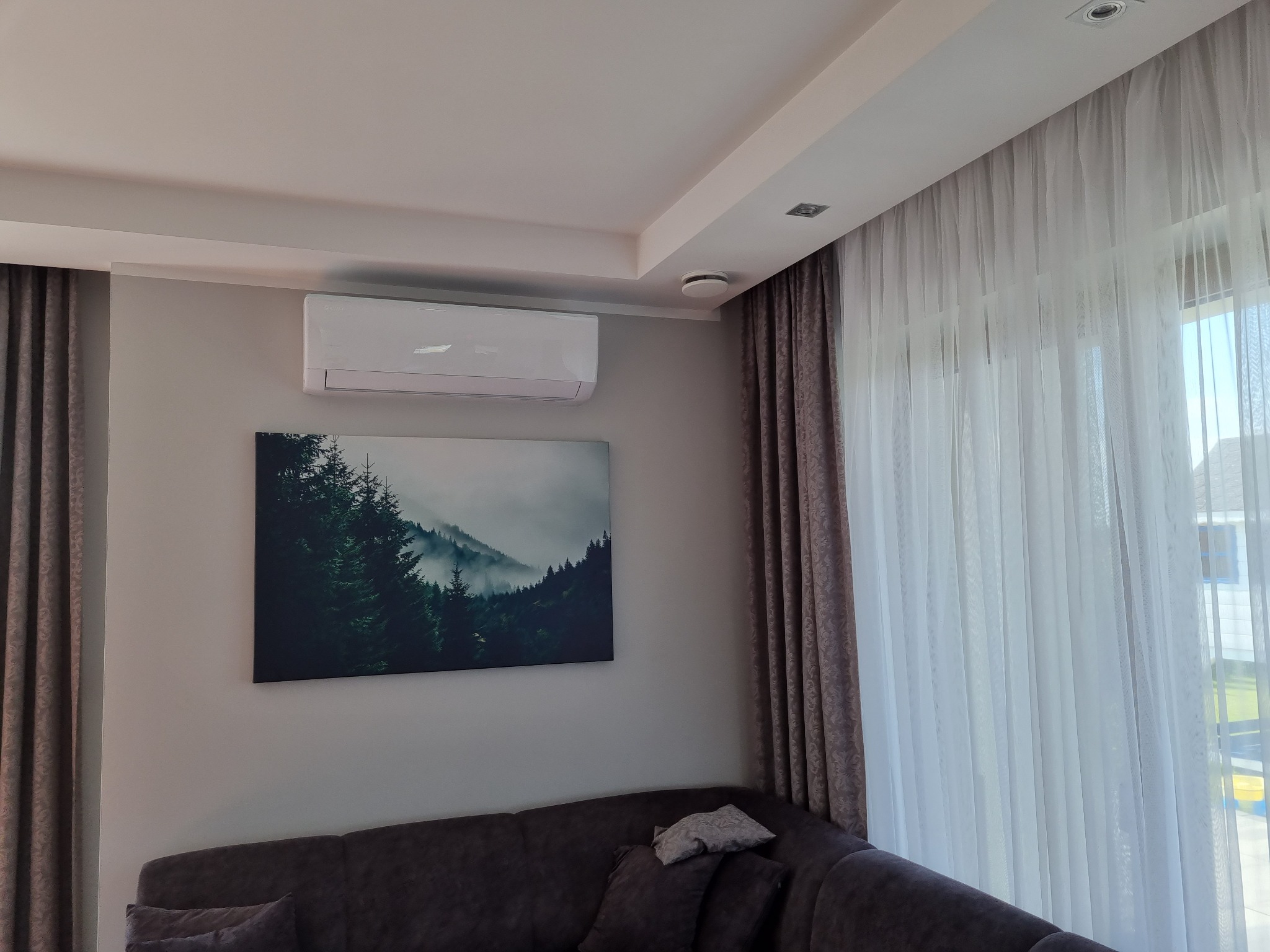 Nowoczesny klimatyzator ścienny zamontowany w salonie nad obrazem z motywem górskiego lasu we mgle, obok okna z firanami i zasłonami.