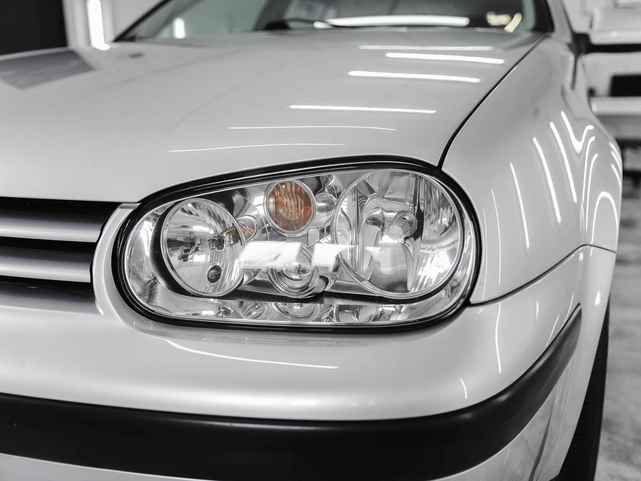 Błyszczący, srebrny samochód z bliska, reflektor odbija światła warsztatu, widoczna czysta powierzchnia lakieru po umyciu.
