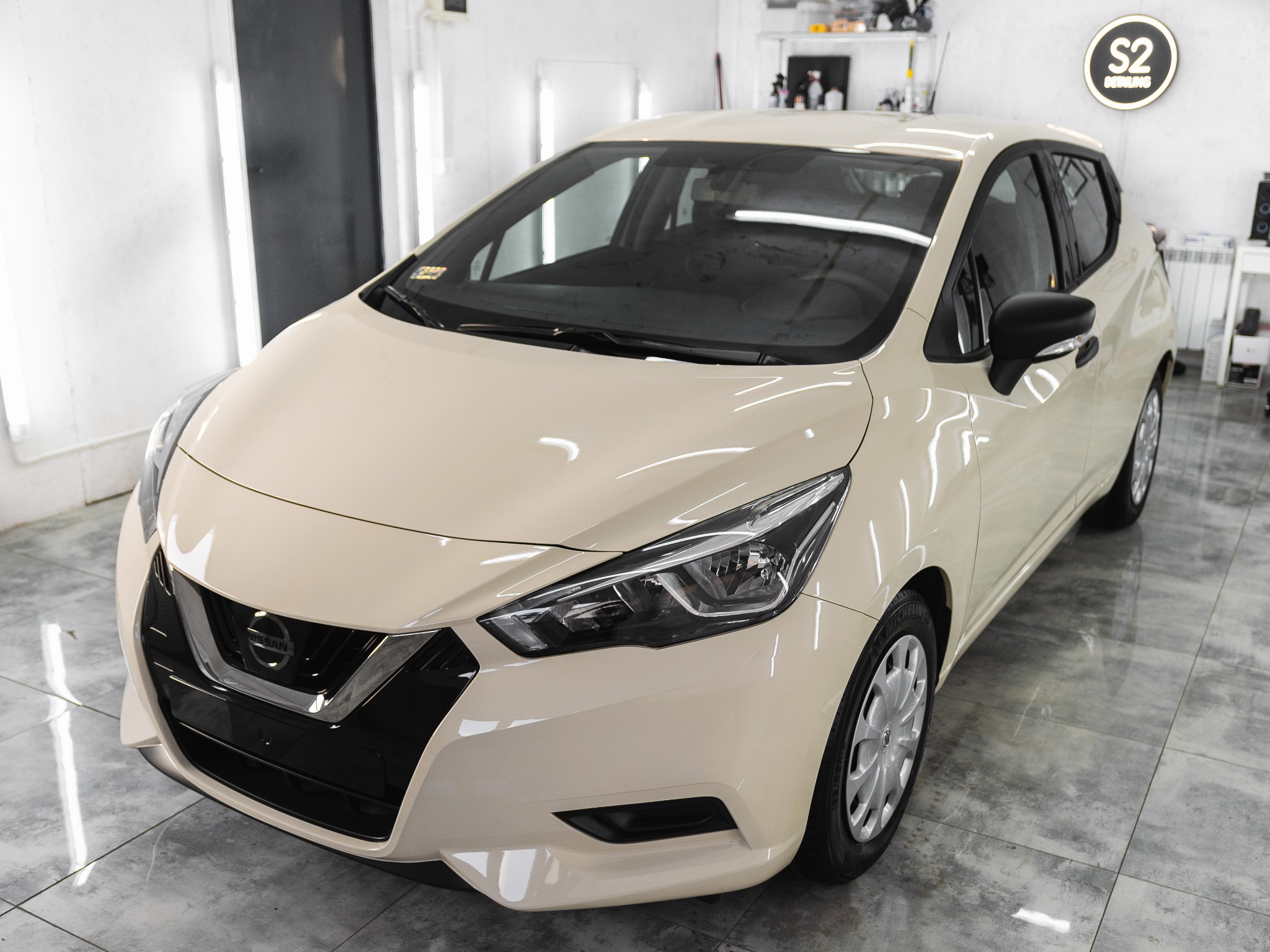 Beżowy Nissan Micra po umyciu, stojący w jasnym pomieszczeniu myjni z błyszczącą podłogą, widoczne logo firmy detailingowej na ścianie.